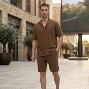 May include: A brown linen shirt and shorts set. The short-sleeved shirt has a button-down front and a chest pocket. The shorts are knee-length. The outfit is paired with white sneakers and a silver watch. The model is standing on a paved walkway.