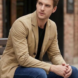 A man in a tan jacket sits on a bench, looking thoughtful.