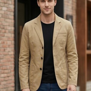 A man wearing a tan blazer jacket, black shirt, and blue jeans stands in front of a brick building.