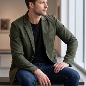 A man in a green blazer and blue jeans sits on a bench, looking off to the side with a thoughtful expression.