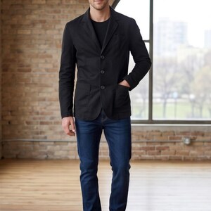 A man wearing a black blazer, blue jeans, and black shoes stands in a room with a brick wall and a large window.
