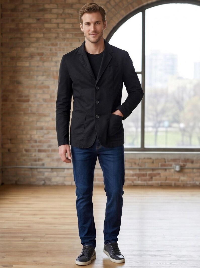 A man wearing a black blazer, blue jeans, and black shoes stands in a room with a brick wall and a large window.