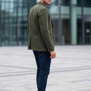 A man wearing a green blazer and dark jeans stands in front of a modern glass building.
