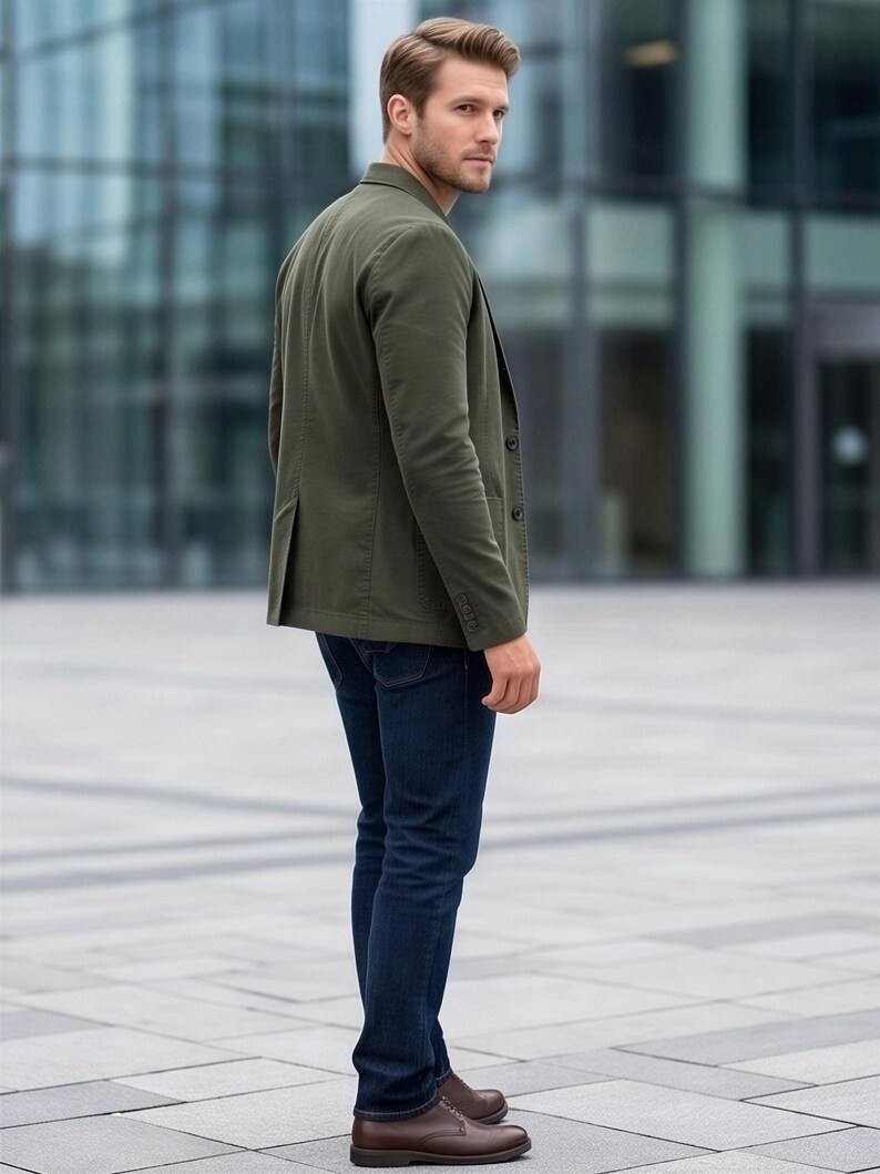A man wearing a green blazer and dark jeans stands in front of a modern glass building.