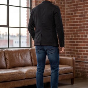 A man wearing a black blazer and jeans stands in a room with a large window and a brown leather couch.