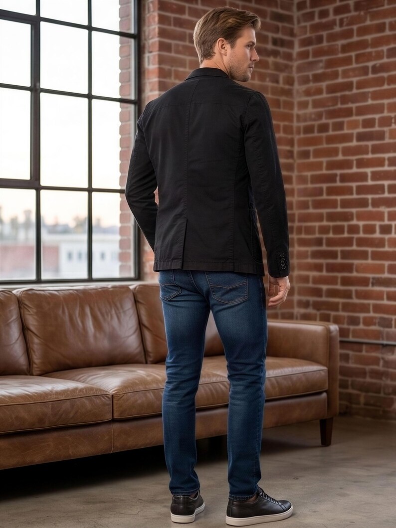 A man wearing a black blazer and jeans stands in a room with a large window and a brown leather couch.