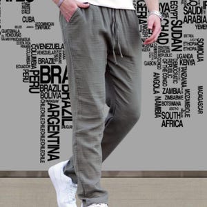 May include: Olive green linen pants with a drawstring waist and pockets. The pants are paired with white sneakers and a white t-shirt. The background features a world map graphic.