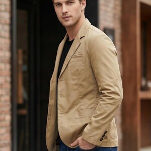 A man wearing a tan blazer and blue jeans stands in front of a brick building.