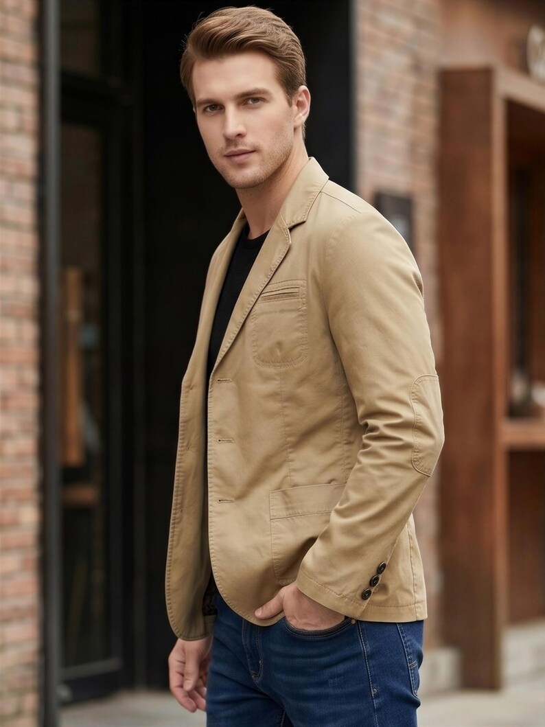 A man wearing a tan blazer and blue jeans stands in front of a brick building.