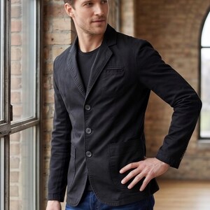 A man wearing a black blazer stands in a room with brick walls and a window.