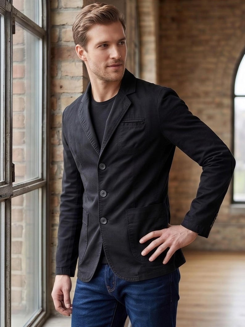 A man wearing a black blazer stands in a room with brick walls and a window.