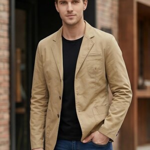 A man wearing a tan blazer and black shirt stands in front of a brick building.