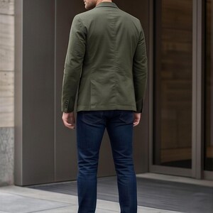 A man wearing a green blazer and blue jeans stands in front of a building.