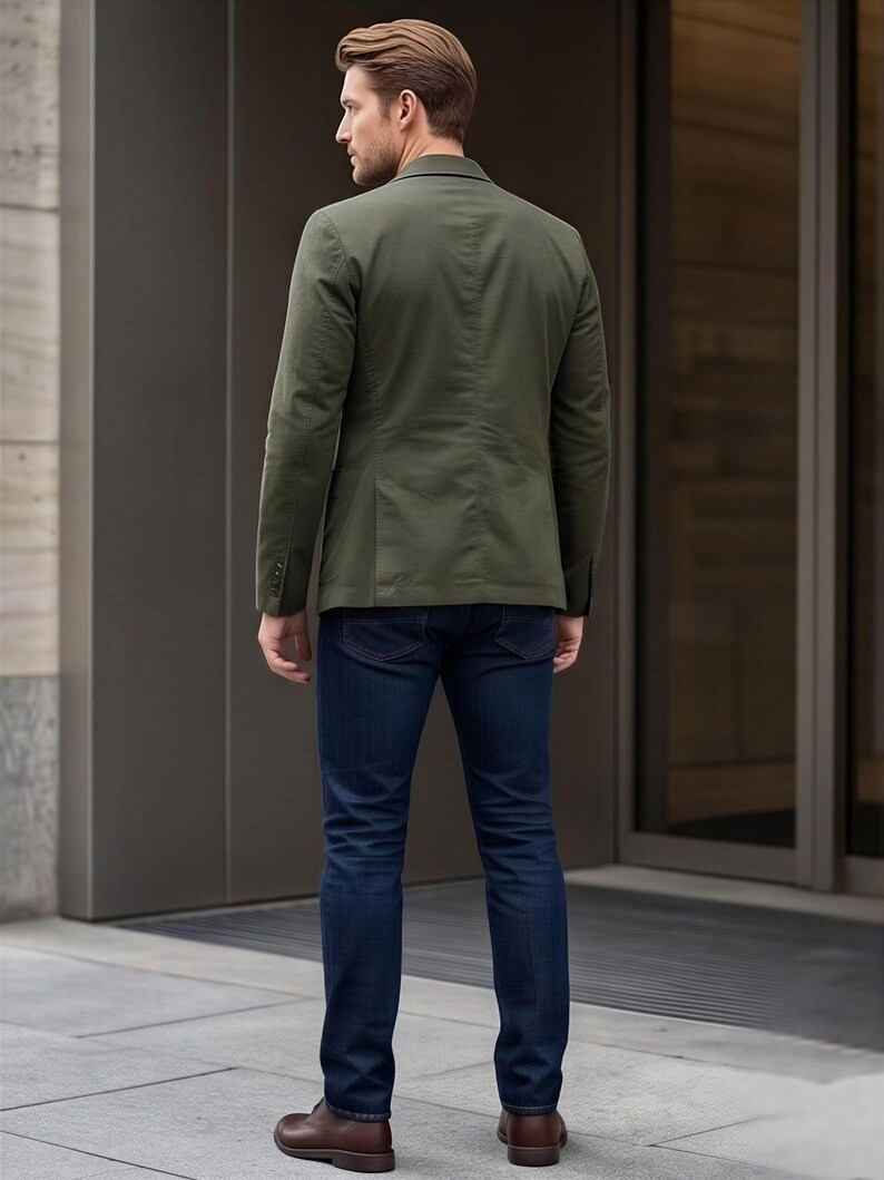 A man wearing a green blazer and blue jeans stands in front of a building.