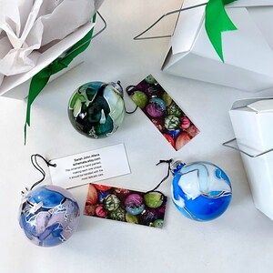 May include: Three hand-painted glass ornaments with unique designs. The ornaments are blue, green, and white with swirls and patterns. The ornaments are on a white surface with a tag that reads "Sarah John Afana schemata.etsy.com This ornament is hand painted making each one unique It should be handled with the most delicate care"