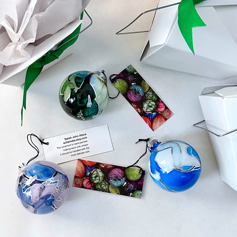 May include: Three hand-painted glass ornaments with unique designs. The ornaments are blue, green, and white with swirls and patterns. The ornaments are on a white surface with a tag that reads "Sarah John Afana schemata.etsy.com This ornament is hand painted making each one unique It should be handled with the most delicate care"