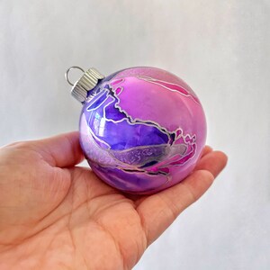 May include: A round glass Christmas ornament with a pink and purple abstract design. The ornament has silver metallic accents.