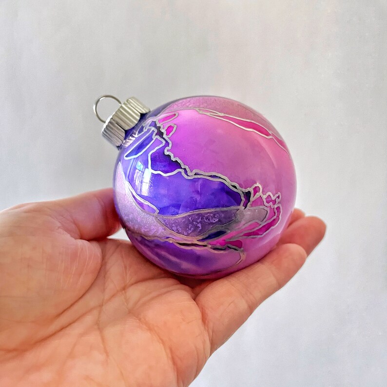 May include: A round glass Christmas ornament with a pink and purple abstract design. The ornament has silver metallic accents.