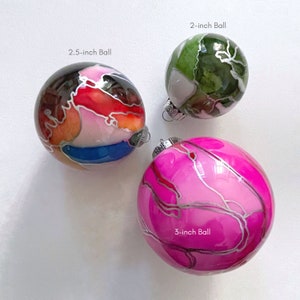May include: Three glass ornaments with different abstract designs. The ornaments are 2, 2.5, and 3 inches in diameter. The 2-inch ornament is green with white and brown accents. The 2.5-inch ornament is red, orange, white, and blue with silver accents. The 3-inch ornament is pink with silver accents.