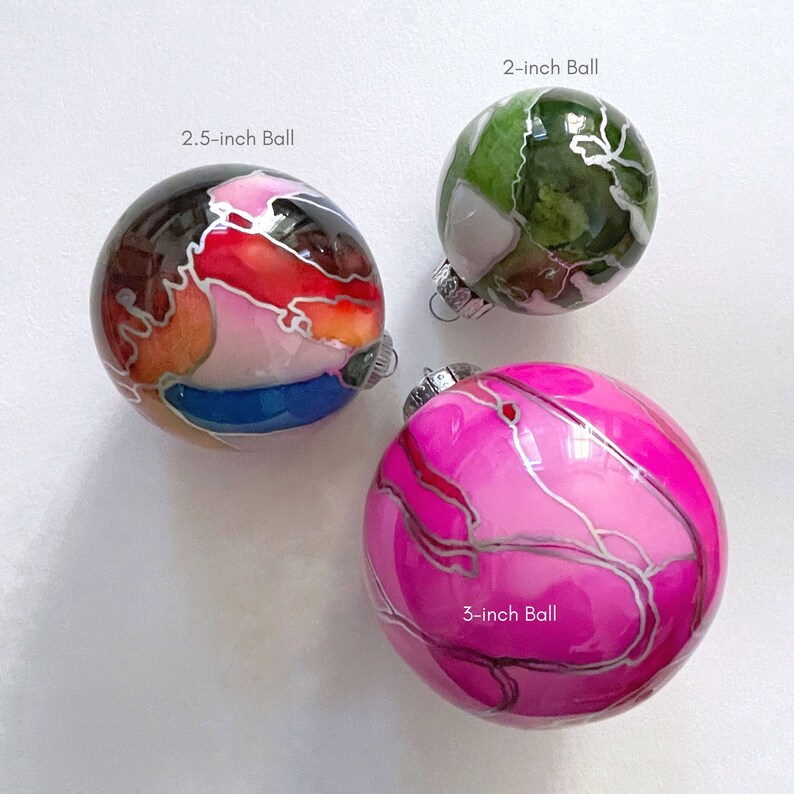May include: Three glass ornaments with different abstract designs. The ornaments are 2, 2.5, and 3 inches in diameter. The 2-inch ornament is green with white and brown accents. The 2.5-inch ornament is red, orange, white, and blue with silver accents. The 3-inch ornament is pink with silver accents.
