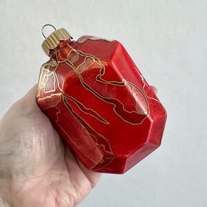 Op de afbeelding: Een facetgeslepen, rode glazen kerstbal met goudkleurige, abstracte ontwerpen. De ornament heeft een goudkleurige metalen dop en een lus om op te hangen. De ornament wordt in een hand gehouden.