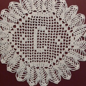 May include: A handmade, off-white crochet doily. The circular doily features an openwork design in the center and a scalloped edge. The intricate pattern is visible against a dark red background. This decorative item is suitable for home decor.