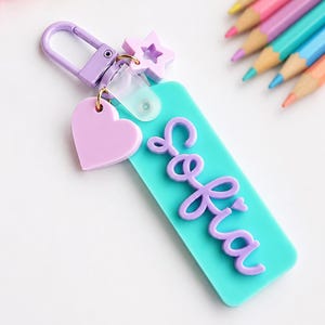 Puede incluir: Un llavero rectangular de acr&iacute;lico turquesa con el nombre Sofia en letras cursivas moradas en relieve. El llavero incluye un cierre de mosquet&oacute;n morado, un peque&ntilde;o dije de estrella morada y un dije de coraz&oacute;n morado claro.