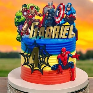 May include: A multi-tiered cake decorated with a Spiderman figure, a web, and a gold number 4. The cake is topped with a gold name banner that reads "GABRIEL" and several superhero figures including Hulk, Iron Man, Thor, and Captain America. The cake is on a silver cake stand with a crystal-like trim.