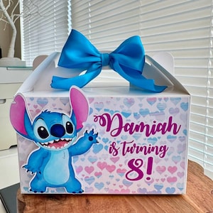May include: A white favor box with a blue bow and a blue Stitch character from the Disney movie Lilo & Stitch. The box has a pink and white heart pattern and the text "Damiah is turning 8!"