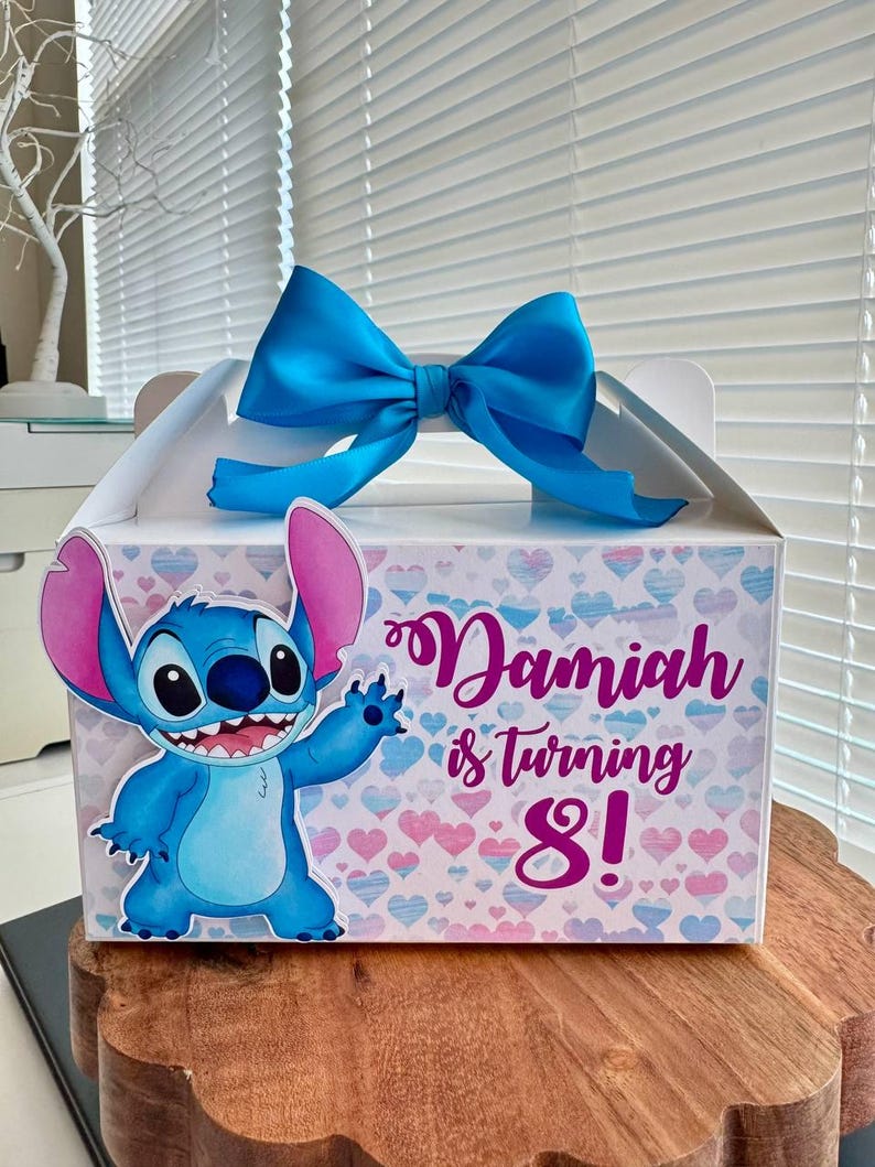 May include: A white favor box with a blue bow and a blue Stitch character from the Disney movie Lilo & Stitch. The box has a pink and white heart pattern and the text "Damiah is turning 8!"