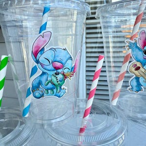 May include: Four clear plastic cups with lids and straws. Each cup has a colorful cartoon sticker of Stitch from Lilo & Stitch. The stickers feature Stitch holding a small plush toy, playing a ukulele, and smiling.