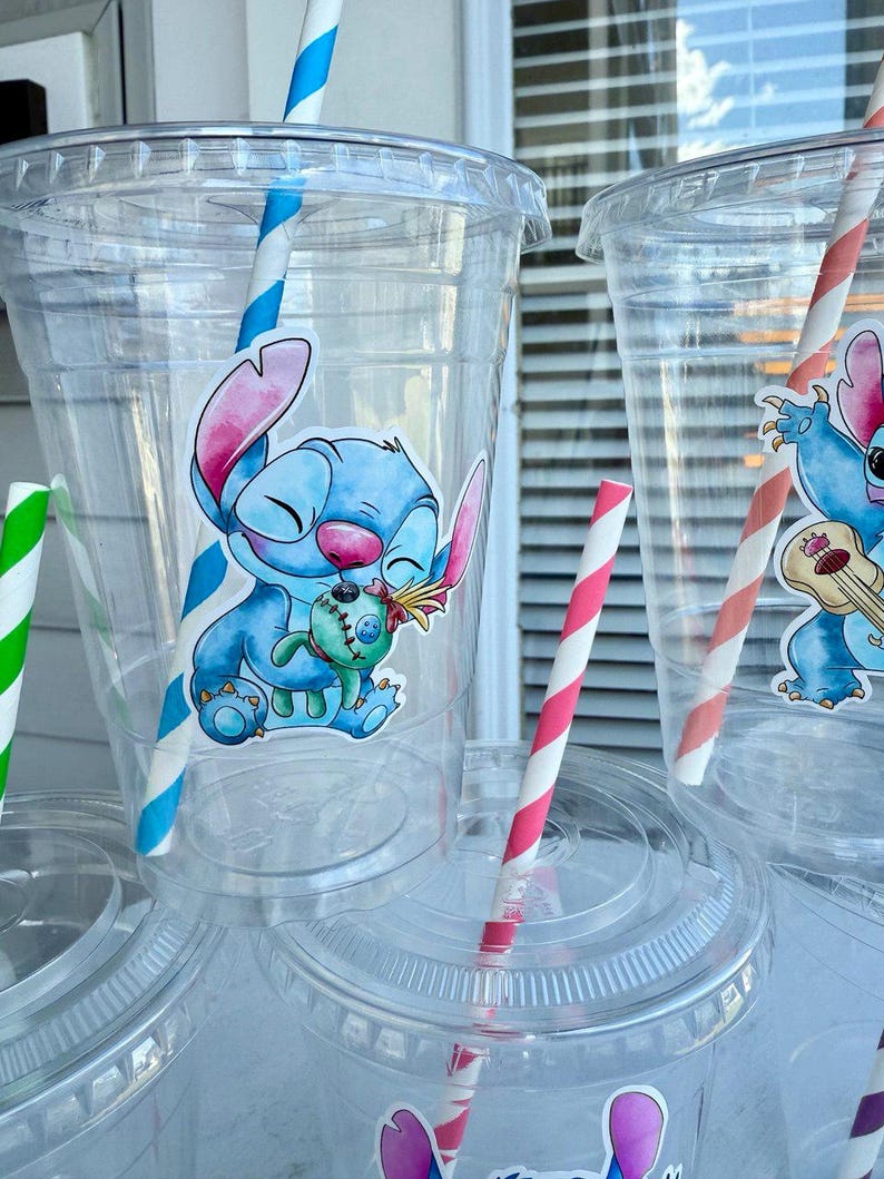 May include: Four clear plastic cups with lids and straws. Each cup has a colorful cartoon sticker of Stitch from Lilo & Stitch. The stickers feature Stitch holding a small plush toy, playing a ukulele, and smiling.