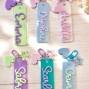 Puede incluir: Seis llaveros rectangulares de acr&iacute;lico con nombres en relieve en letra cursiva: Emma, Daphne, Arlette, Sabrina, Scarlett y Crimson. Cada llavero incluye un cierre de mosquet&oacute;n, un dije de coraz&oacute;n y uno de estrella en tonos pastel.