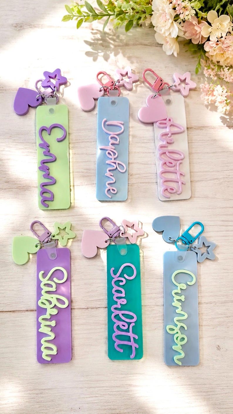 Puede incluir: Seis llaveros rectangulares de acr&iacute;lico con nombres en relieve en letra cursiva: Emma, Daphne, Arlette, Sabrina, Scarlett y Crimson. Cada llavero incluye un cierre de mosquet&oacute;n, un dije de coraz&oacute;n y uno de estrella en tonos pastel.