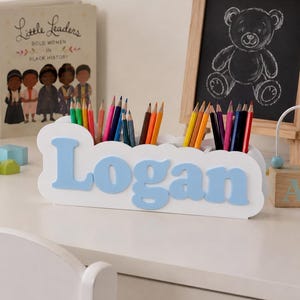 May include: A white wooden pencil holder with a scalloped edge and the name Logan in raised light blue letters. The organizer features multiple compartments filled with an assortment of colored pencils in various shades including red, blue, and yellow.