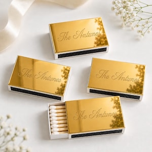 May include: Four rectangular matchboxes with a reflective gold metallic finish. Each box features the engraved text The Antunes in a cursive script. One box is partially open, revealing wooden matches with white tips. The boxes have black striking strips.