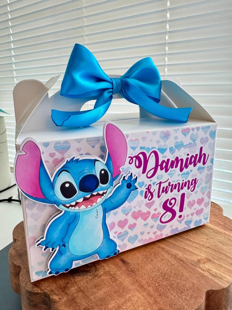 May include: A blue and pink party favor box with a blue bow. The box features a cartoon Stitch character and the text "Damiah is turning 8!"