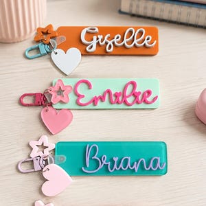 Puede incluir: Cuatro llaveros rectangulares de acr&iacute;lico con nombres personalizados en letras cursivas en relieve. Cada llavero incluye un cierre met&aacute;lico, un dije en forma de estrella y un dije en forma de coraz&oacute;n, en colores naranja, verde menta, turquesa y rosa.