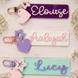 Puede incluir: Tres llaveros rectangulares con dijes de estrella y coraz&oacute;n. El llavero superior es morado con el nombre Eloise en blanco. El del medio es blanco con el nombre Aaliyah en rosa. El inferior es verde claro con el nombre Lucy en morado.