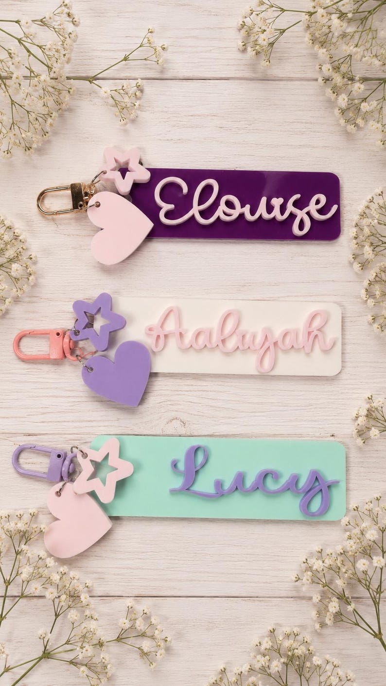 Puede incluir: Tres llaveros rectangulares con dijes de estrella y coraz&oacute;n. El llavero superior es morado con el nombre Eloise en blanco. El del medio es blanco con el nombre Aaliyah en rosa. El inferior es verde claro con el nombre Lucy en morado.