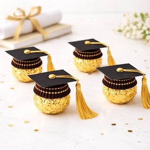 May include: Four graduation-themed treats. Each features a gold-wrapped candy base, topped with a black mortarboard, gold tassel, and a decorative brown and gold trim. A rolled diploma and confetti are in the background.