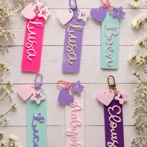 Puede incluir: Seis llaveros rectangulares de acr&iacute;lico con cierres de mosquet&oacute;n de metal. Cada uno presenta un nombre en letras cursivas en relieve, un dije de coraz&oacute;n y un dije de estrella. Los colores incluyen rosa intenso, lavanda, verde menta y morado.