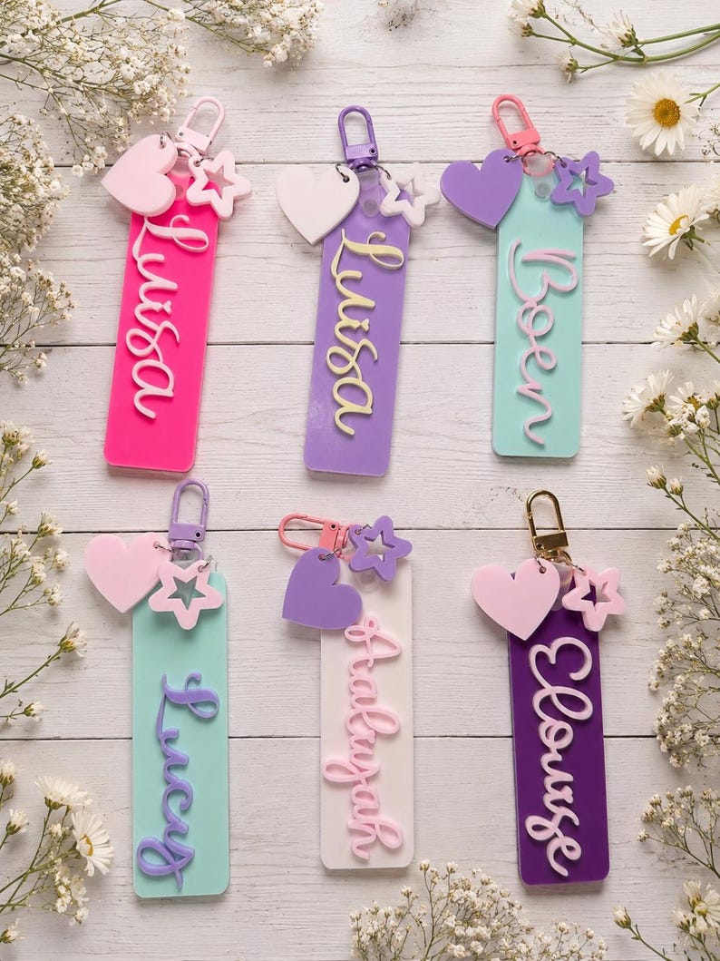Puede incluir: Seis llaveros rectangulares de acr&iacute;lico con cierres de mosquet&oacute;n de metal. Cada uno presenta un nombre en letras cursivas en relieve, un dije de coraz&oacute;n y un dije de estrella. Los colores incluyen rosa intenso, lavanda, verde menta y morado.