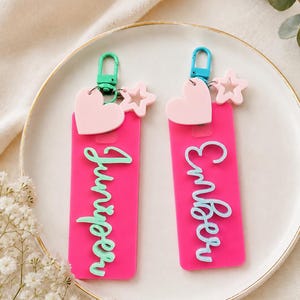 Puede incluir: Dos llaveros rectangulares de acr&iacute;lico rosa intenso con los nombres Juniper y Ember en letras cursivas en relieve. Cada llavero incluye un dije de coraz&oacute;n rosa claro, una peque&ntilde;a estrella y un cierre de mosquet&oacute;n de pl&aacute;stico colorido.