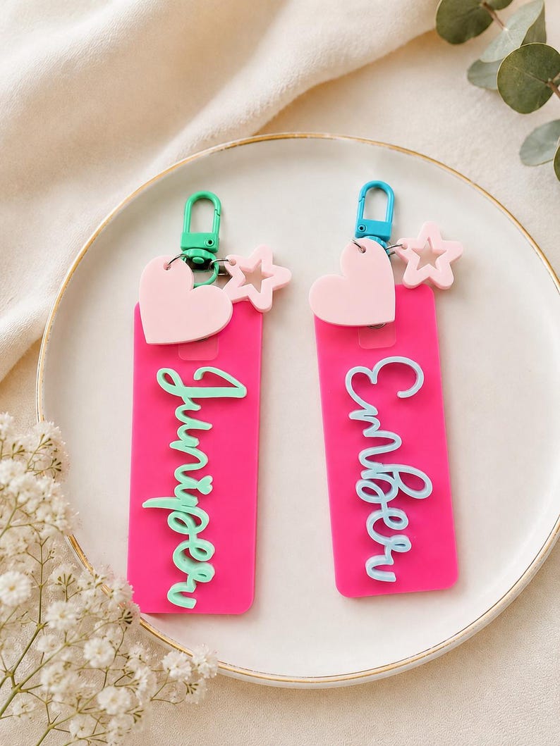 Puede incluir: Dos llaveros rectangulares de acr&iacute;lico rosa intenso con los nombres Juniper y Ember en letras cursivas en relieve. Cada llavero incluye un dije de coraz&oacute;n rosa claro, una peque&ntilde;a estrella y un cierre de mosquet&oacute;n de pl&aacute;stico colorido.