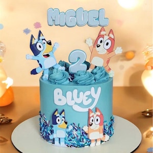 May include: A light blue cake decorated with Bluey characters and the name "Miguel". The cake has a "2" on top, with the word "Bluey" written on the side. The cake is on a white cake board.