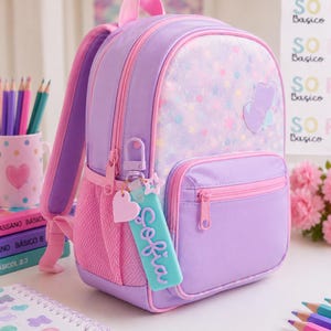 Puede incluir: Una mochila de color lavanda y rosa con un panel frontal estampado de estrellas, un bolsillo frontal con cremallera y bolsillos laterales de malla. Un llavero rectangular azul claro con el nombre Sofia y un peque&ntilde;o coraz&oacute;n rosa cuelga de la cremallera.