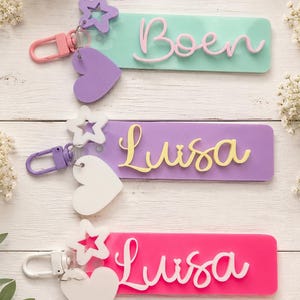 Puede incluir: Tres llaveros rectangulares de acr&iacute;lico con dijes de estrella y coraz&oacute;n. El llavero superior es verde menta con el nombre Boen en rosa. El del medio es morado con el nombre Luisa en amarillo. El inferior es rosa intenso con el nombre Luisa en blanco.