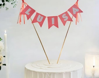 May include: A white cake with a pink and white banner that says "LUISA" on top. The banner is attached to two wooden sticks.