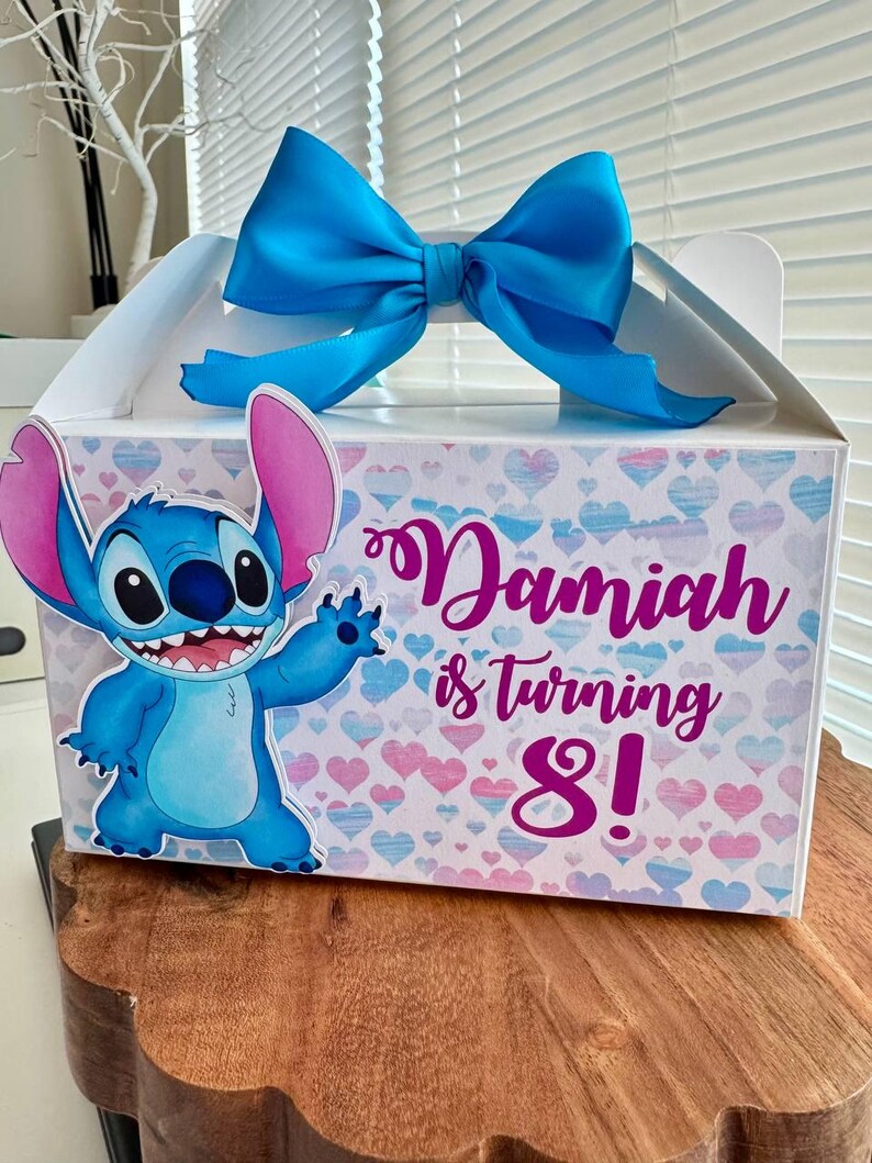 May include: A blue and pink party favor box with a blue bow. The box features a cartoon Stitch character and the text "Damiah is turning 8!"